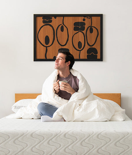 Man holding mug and wrapped in washable wool comforter sits on top of a Quail Responsive Foam Mattress